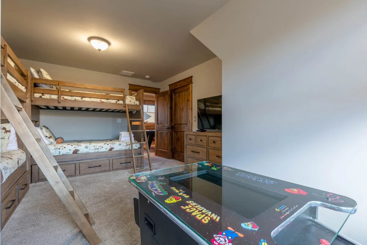 A cozy corner of the loft or bunk area, featuring classic wooden bunk beds and a dedicated arcade game table. A TV sits nearby, enhancing the recreational aspect, while wood tones and soft lighting create a welcoming feel perfect for late-night gaming and sibling bonding.