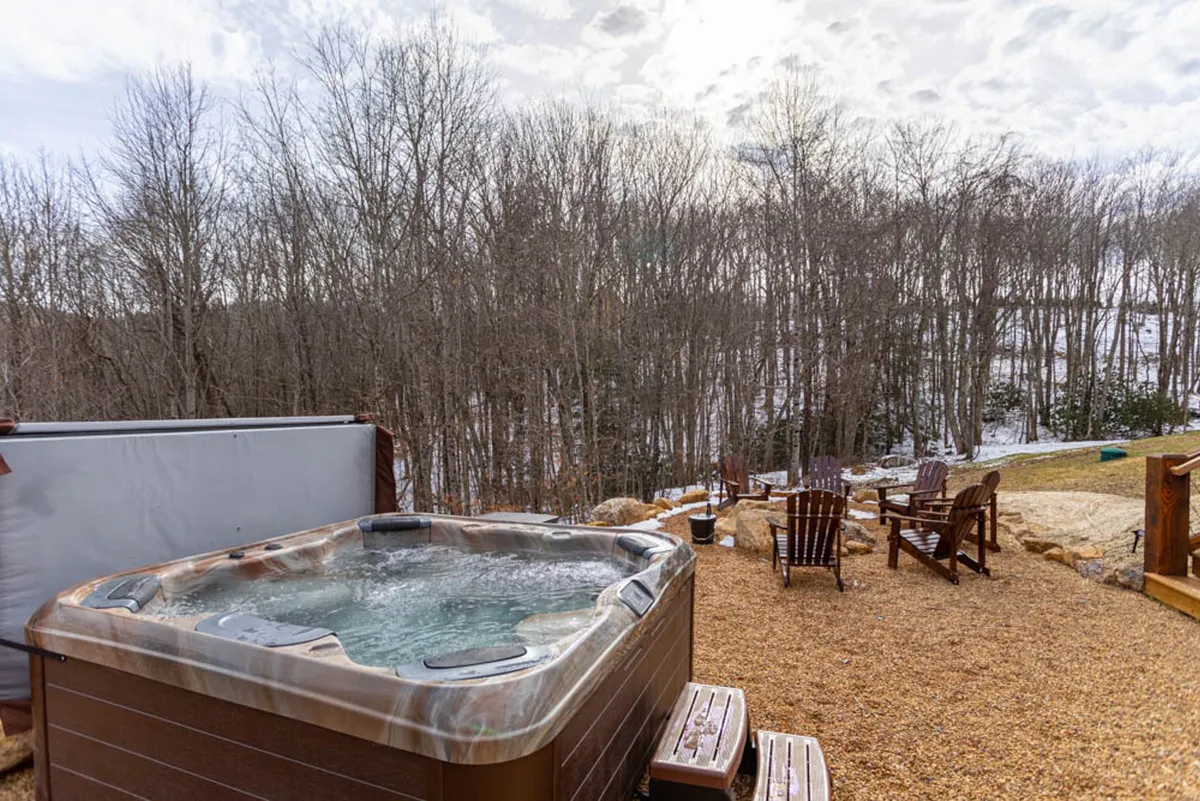 An inviting outdoor scene showcasing a bubbling hot tub, Adirondack chairs, a gravel patio, and a forested backdrop. The set-up suggests relaxation and quality family time spent under the open sky, with the fire pit area hinting at evening marshmallow roasting and social gatherings.