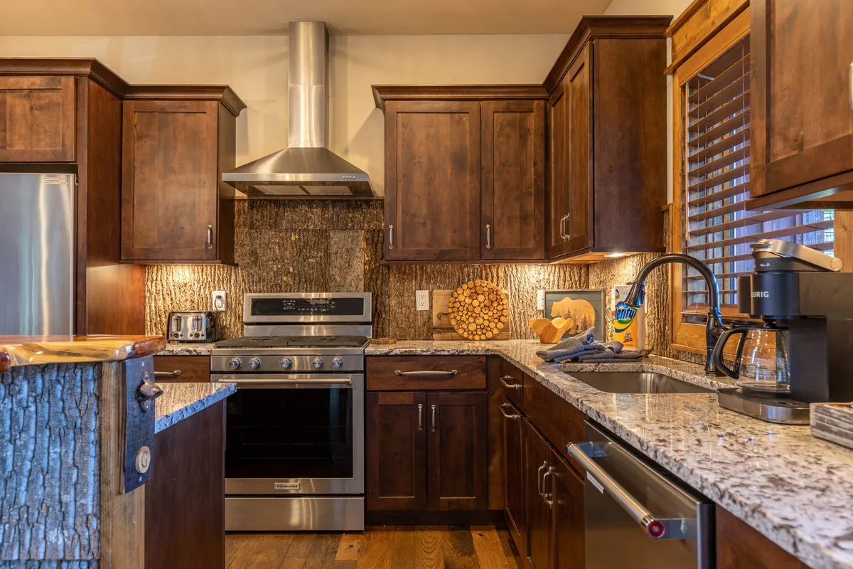 A stylish kitchen vignette featuring granite countertops, wooden cabinetry, stainless steel appliances, a textured backsplash, and a decorative wooden bowl. The modern yet cozy design invites both culinary creativity and socializing.