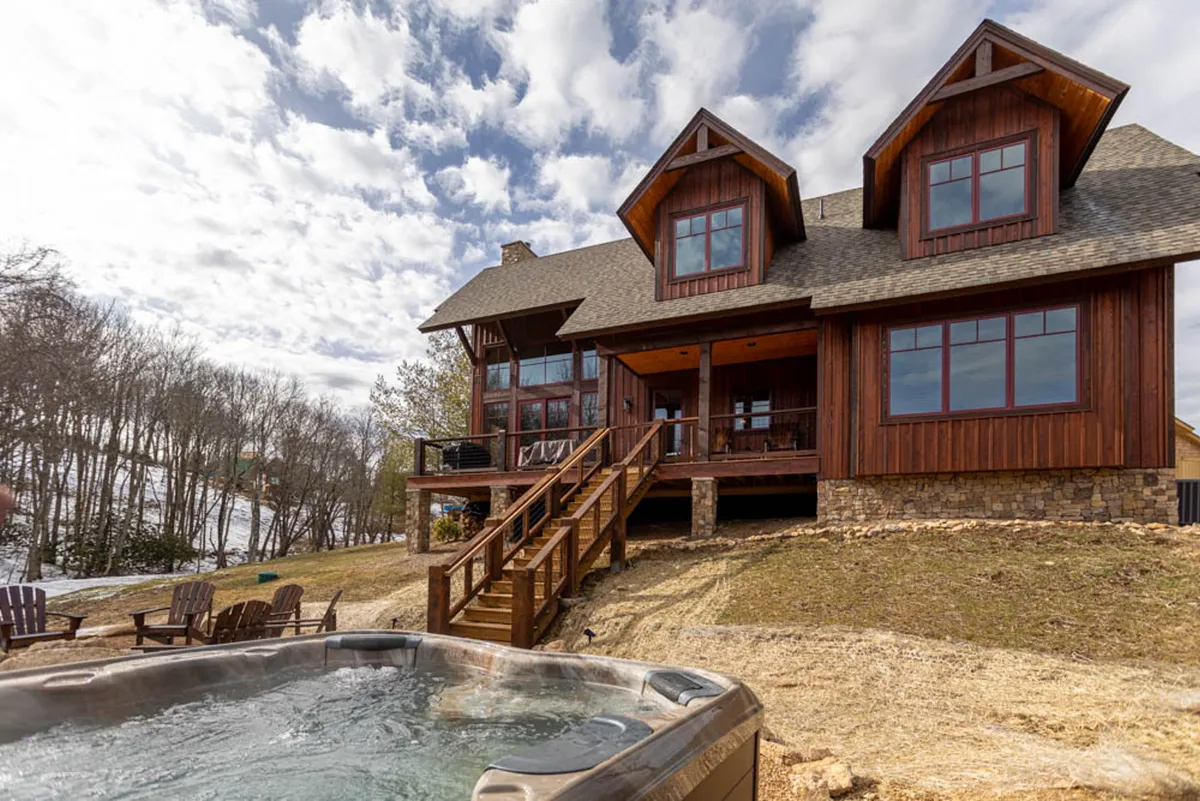 The hero image displays the exterior of Little Cubs Cabin, showcasing its inviting wooden deck with a bubbling hot tub, expansive large windows that reflect the surrounding nature, and a landscaped yard perfect for outdoor dining and gatherings. The scene evokes a sense of serene mountain living with opportunities for stargazing, sunset views, and relaxing in comfort.