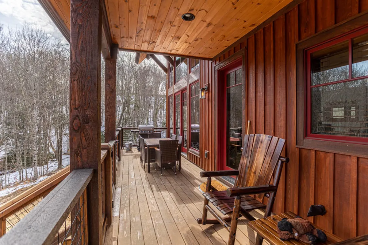 An inviting outdoor deck featuring a wooden rocking chair and a dining set, bordered by a wooden railing and surrounded by lush, mature trees. The atmosphere is cozy and welcoming, seamlessly blending relaxation with the outdoors.