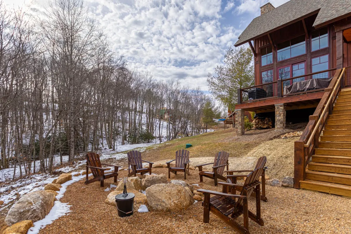 A cozy outdoor space with a fire pit surrounded by Adirondack chairs, wooden steps leading up to the home’s facade, and lush woodland surroundings. The area invites friendly gatherings and relaxation, embodying the spirit of roasting marshmallows and intimate evenings with family or friends.