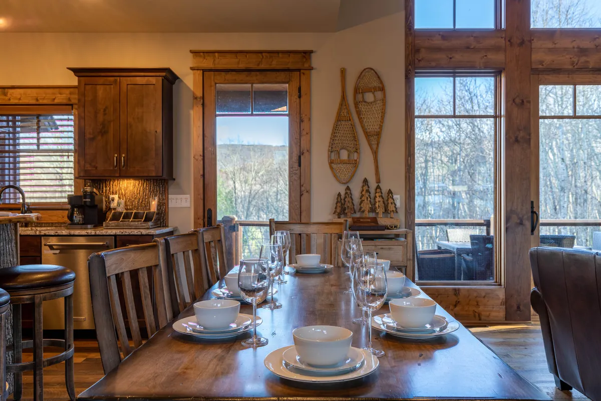A warmly lit dining area with a rustic wooden table set for a meal, flanked by large windows that let in natural light and offer glimpses of the outdoors. Decor elements like snowshoes on the wall and sturdy wooden cabinetry evoke a cozy mountain cabin vibe.