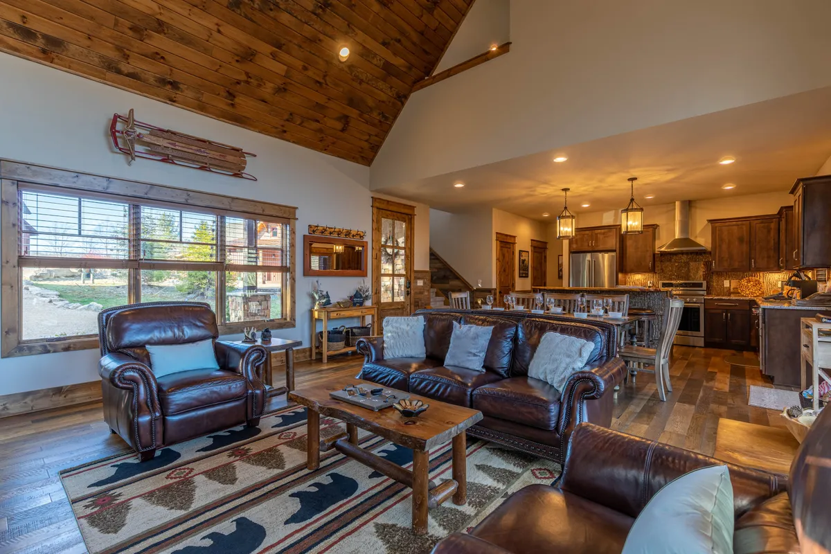 A bright, open-concept main living space featuring plush leather sofas gathered around a central fireplace, a large wooden dining table, and a visible kitchen area. Natural light streams in through expansive windows, and the cozy rug with bear motifs adds a touch of mountain charm. The kitchen is seamlessly integrated, perfect for socializing while cooking.